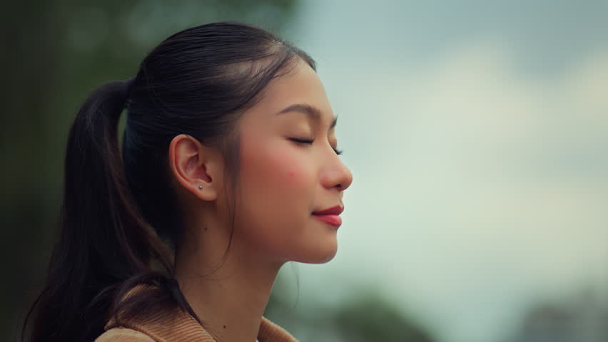 Asian woman breathing deeply and opening eyes with calm smile, young female enjoying fresh air outdoors, concept of mindfulness, mental health, relaxation, and self care in peaceful environment.