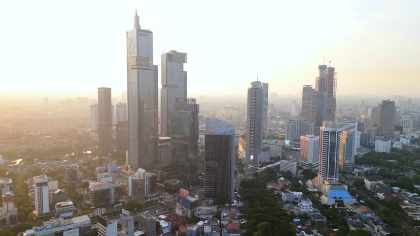 Aerial drone view of Jakarta skyline at sunset with tall skyscrapers glowing in golden light. Modern cityscape of Indonesia