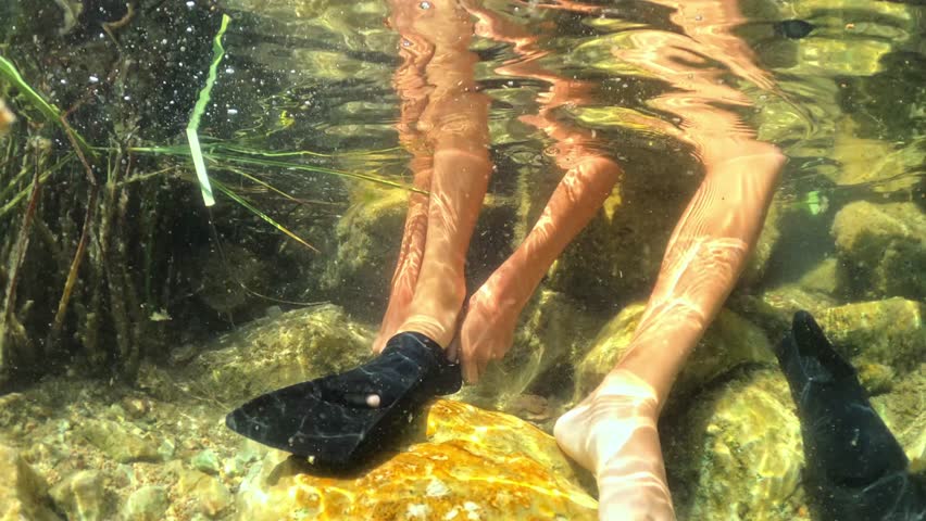 Person perched on rock slipping on black flippers, preparing to snorkel in crystal-clear sunlit water as sunlight creates shimmering caustic patterns on the seabed
