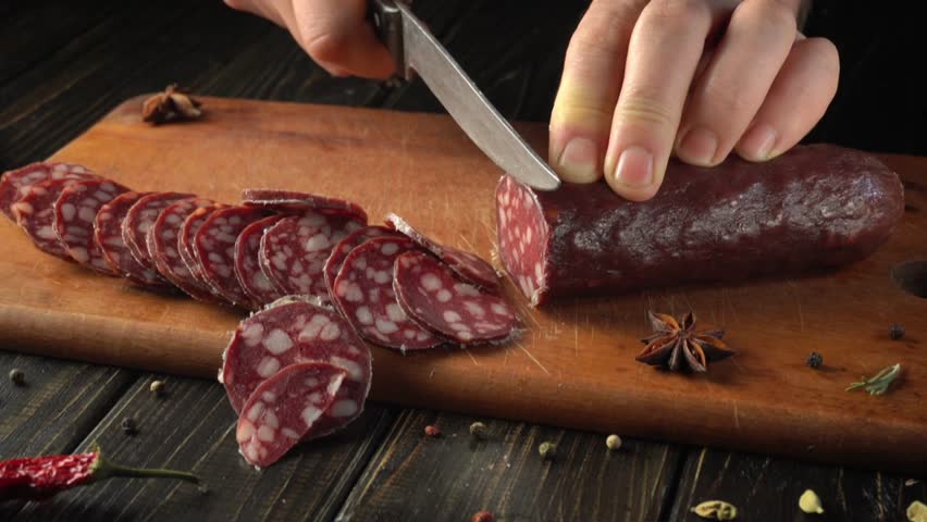 Hands are slicing a cured sausage on a wooden cutting board. Colorful spices are spread across the surface, creating a warm, inviting kitchen atmosphere. The focus is on the precision of cutting.