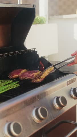 Vertical close-up shot of a beef steak being grilled on an outdoor barbecue, flipped with tongs over open flames, with asparagus cooking on the side