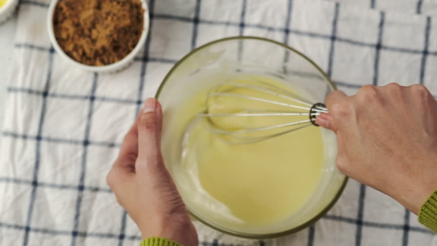 top-down close-up of hands whisking smooth batter in glass bowl on checkered cloth, surrounded by simple baking ingredients; cozy homemade dessert preparation moment
