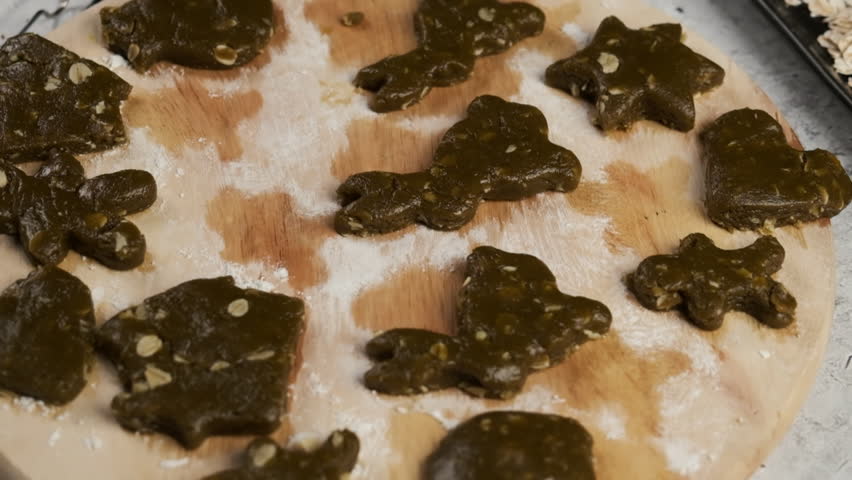 Christmas-themed green cookie dough cut into festive shapes like stars, hearts, and trees, laid out on floured wooden board ready for baking
