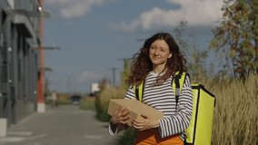 Cheerful woman courier, donning a thermal backpack, stands on a city street next to a sleek building on a sunny day, holding a cardboard package for delivery and smiling directly at the camera - Powered by Shutterstock - Get 15% off with code: PIKWIZARD15