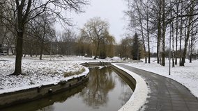 The partially ice-covered canal runs through the park among spruce, poplars and birch trees. The banks are concreted. Snow covers the ground. Nearby there is a tiled path, lampposts and buildings - Powered by Shutterstock - Get 15% off with code: PIKWIZARD15