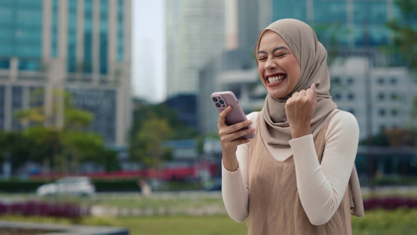 Portrait of surprised young businesswoman wearing hijab reads exciting news on smartphone stands in downtown. Malaysian lady expresses mix of amazement and happiness on city street - Powered by Shutterstock - Get 15% off with code: PIKWIZARD15
