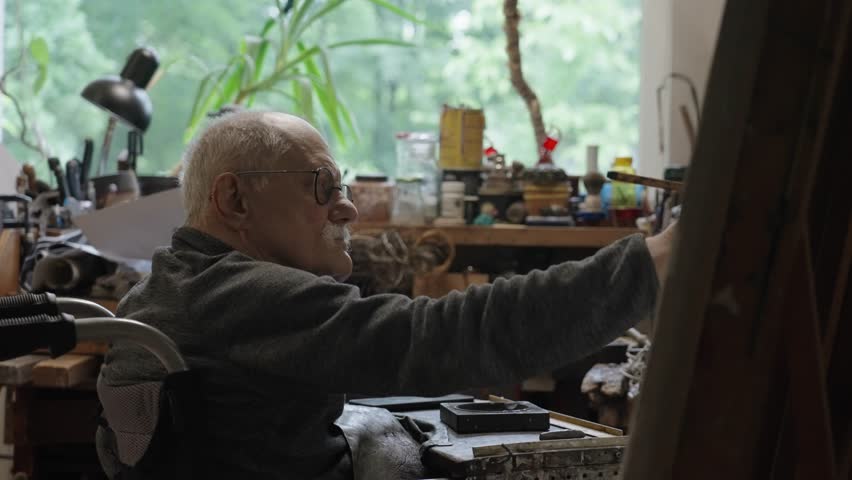 Senior artist on a wheelchair paints an oil painting on canvas in a studio