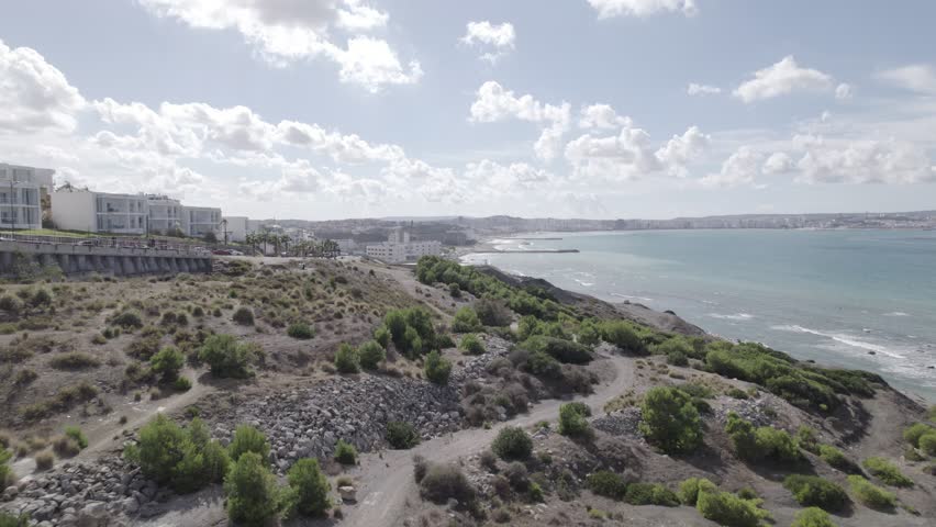 Aerial drone view of a coastal hillside in Morocco, showing rocky terrain, scattered green shrubs, seaside buildings, and the bright blue ocean under a sunny sky.