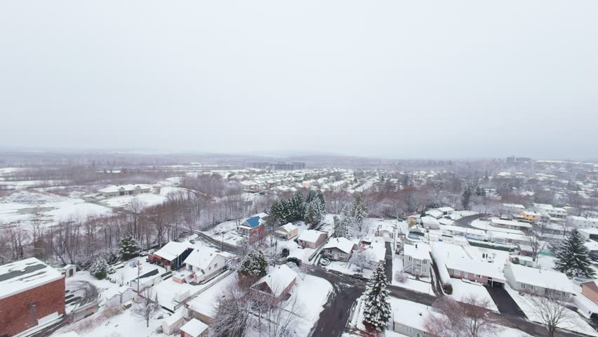 Calm aerial motion above snowy Canadian suburban streets under soft white winter sky and gentle snowfall.