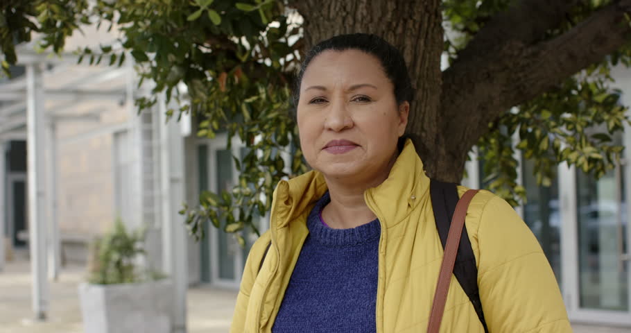 Hispanic woman spotting camera leaning on tree trunk on sidewalk smiling laughing with backpack. Urban, lifestyle, candid, natural, casual, contemporary, expressive