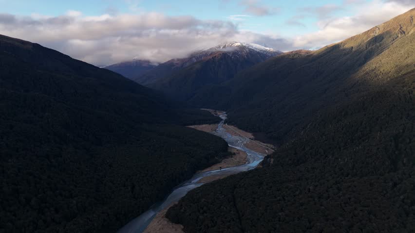 Flowing river between giant mountains and snowy peak at dusk. Aerial wide shot. New Zealand, South Island, Mount Aspiring area.