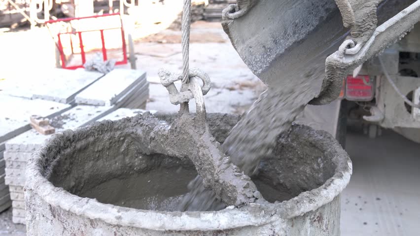 A close-up mixed wet concrete being poured from the chute of a concrete mixer truck into a large, sturdy bucket suspended by a wire cable and chain mechan.