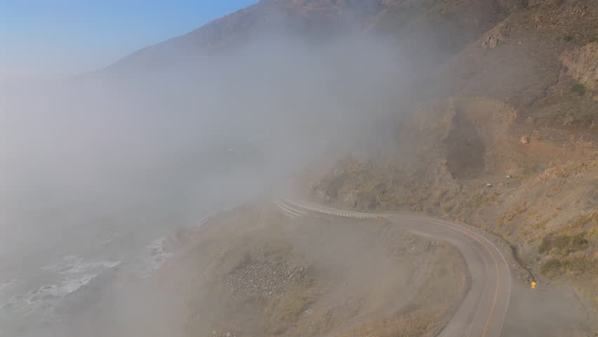 Aerial drone view of Big Sur California with fog drifting over rugged cliffs and the winding coastline under soft daylight, showing the dramatic Pacific scenery for travel and nature themes