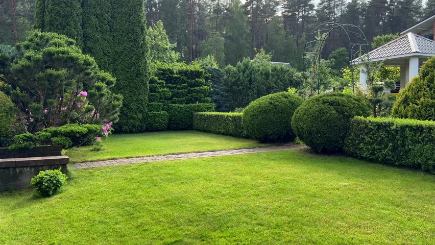 A bright, well-maintained formal garden with a lush green lawn, a brick path, and various manicured evergreen shrubs, including spherical topiary and layered hedges, on a sunny day