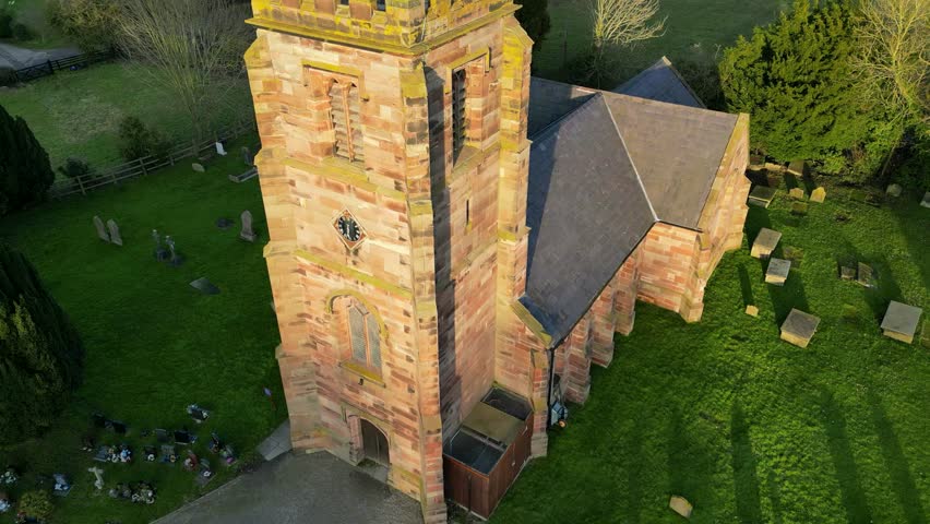 Aerial view of historic stone church with clock tower and surrounding graveyard in rural Hampshire, England, showing warm sunlight over green countryside and traditional village setting.