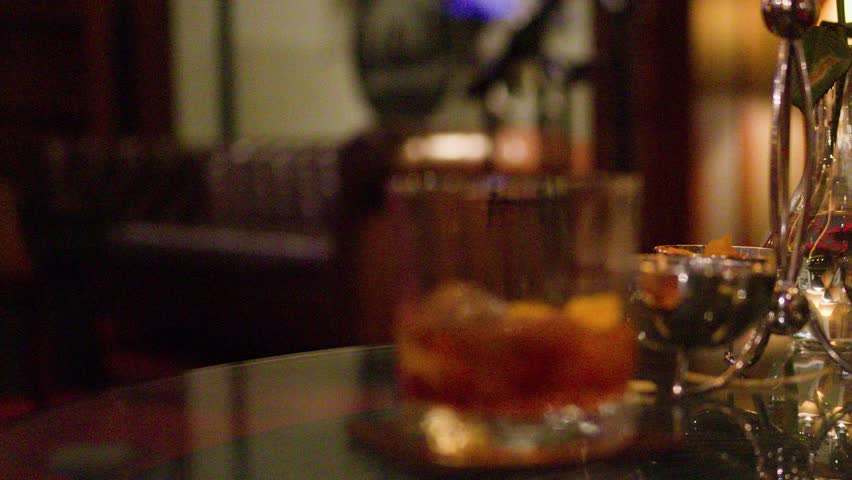 Whiskey glass with ice on reflective bar table, warm lounge lighting, slow focus shift