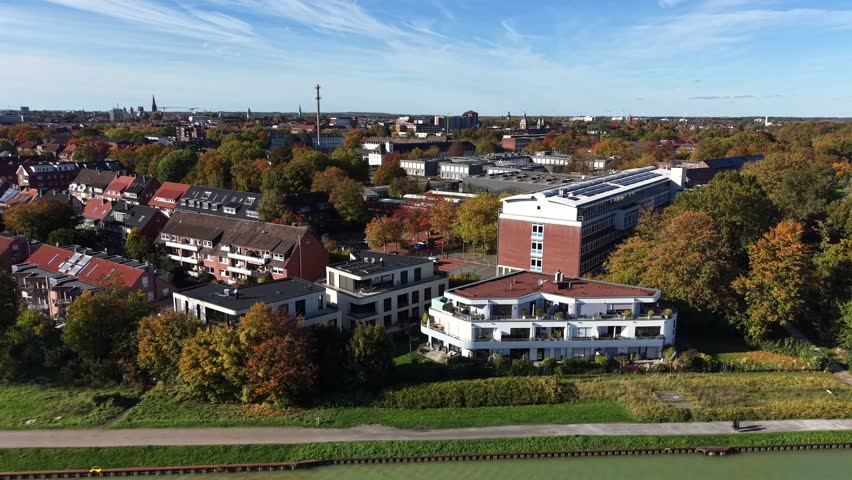 Aerial lateral shot of German city architecture in autumn, with red-roofed houses, modern buildings and colorful fall trees under a clear blue sky. Wide shot. Green river water in foreground.