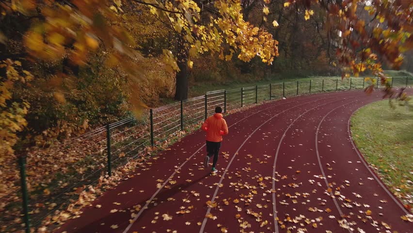 Man running on red track in autumn park. Man in orange sportswear jogging at sport stadium with fallen autumn leaves. Concept of fitness, wellness and outdoor training