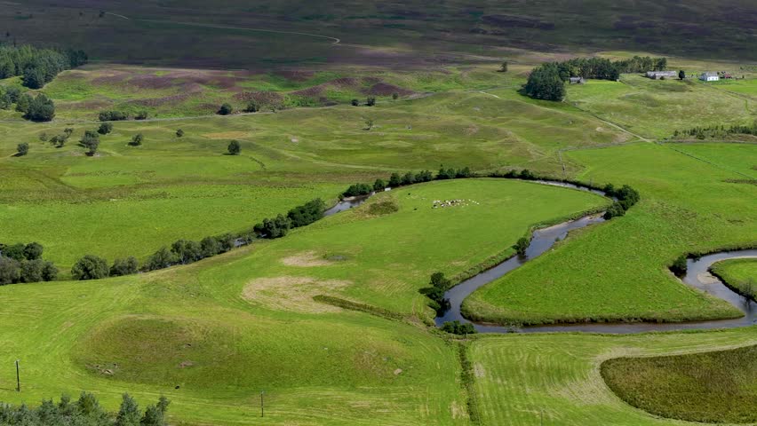 Drone glides above winding river, lush green meadows, distant hills, and scattered trees, daylight