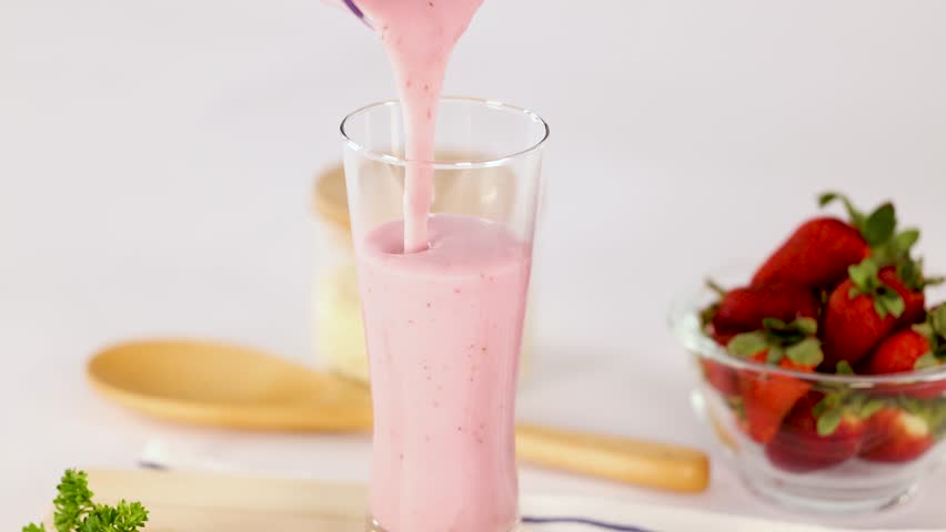 Pink strawberry smoothie poured into glass, surrounded by fresh berries, wooden spoon, bright lighting