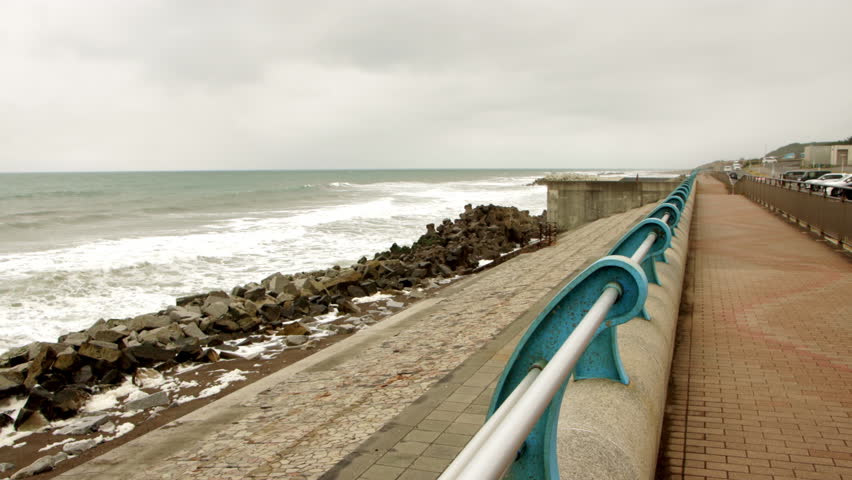 Static wide shot of ocean waves breaking against a rocky seawall beside an empty coastal promenade in Japan on an overcast day