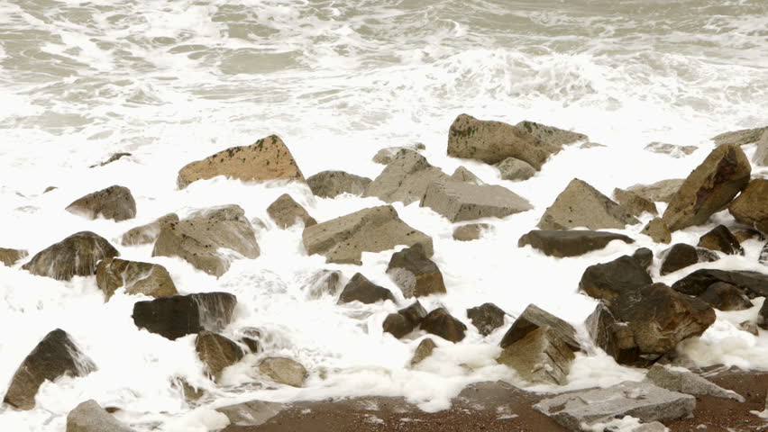 Static shot of ocean waves crashing over large coastal rocks along the shoreline in Japan on an overcast day