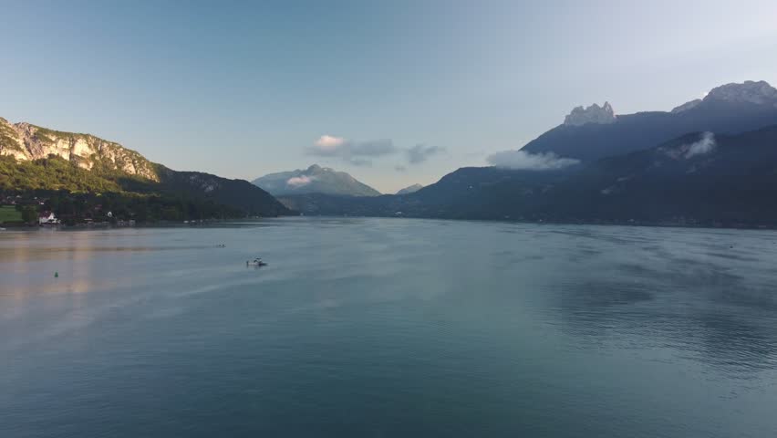 Rotational drone footage of Lake Annecy just after Sunrise. The sun has just appeared over the mountain tops and the odd boat is already on the water. Footage moves to the left. Clouds on the hills.
