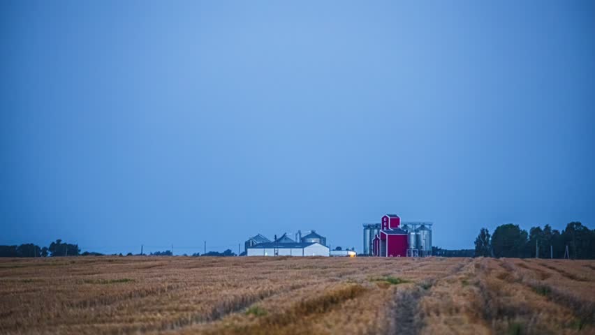 Wide shot of harvested grain field with red silo and farm buildings at dusk