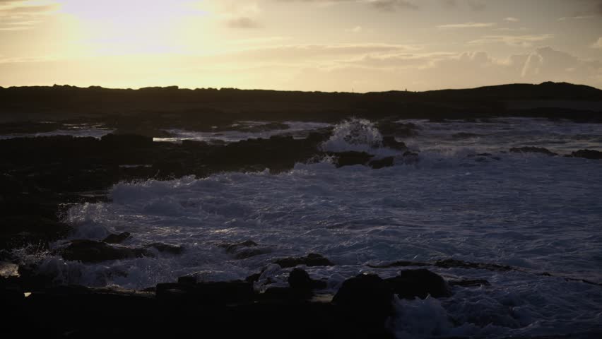Golden light fades over a rugged shoreline where waves collide with black volcanic rocks, capturing the ocean’s power and the tranquil beauty of dusk.