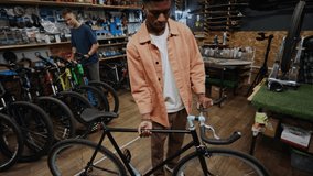 Young Black man working in repair shop, lifting bicycle to check its handlebar and steerer tube, smiling at camera, his Caucasian colleague working in background - Powered by Shutterstock - Get 15% off with code: PIKWIZARD15