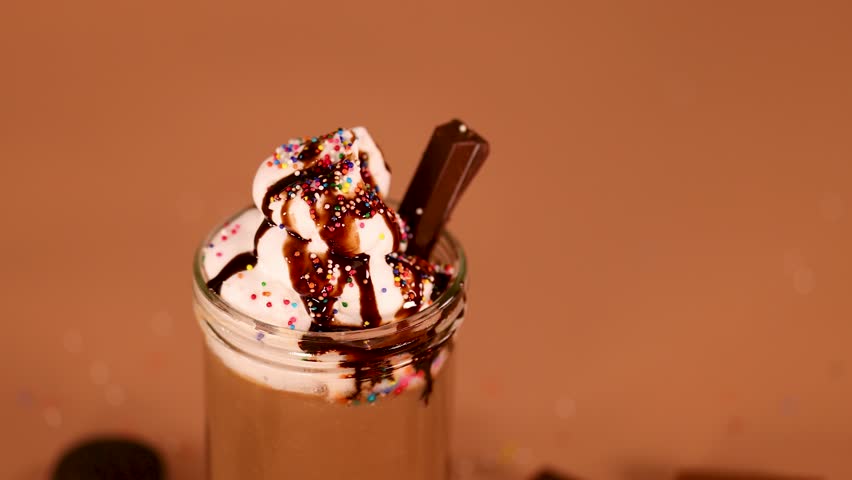 Hand places cherry atop whipped cream, chocolate syrup, and sprinkles on milkshake under warm lighting.