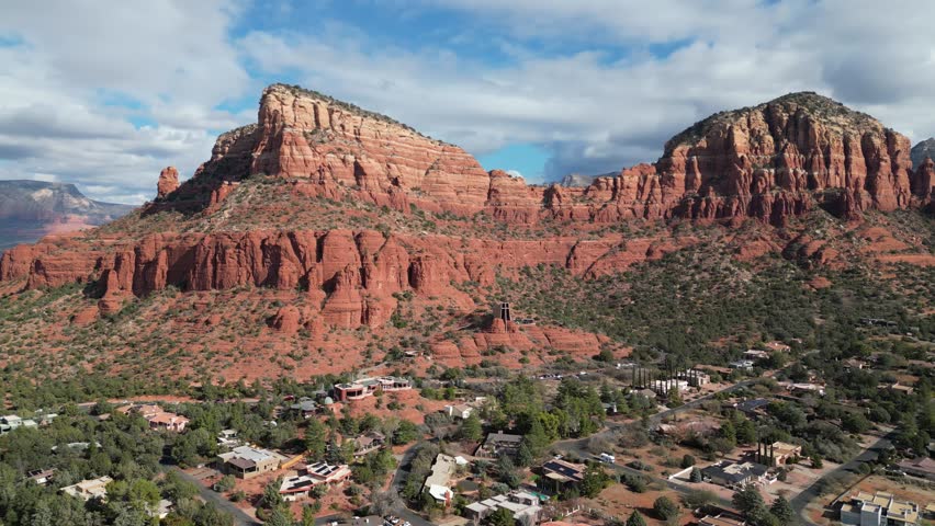 Aerial drone footage of Chapel of the Holy Cross in Sedona, Arizona, showing the modern architectural landmark built into red sandstone cliffs surrounded by desert landscape and scenic red rock views.