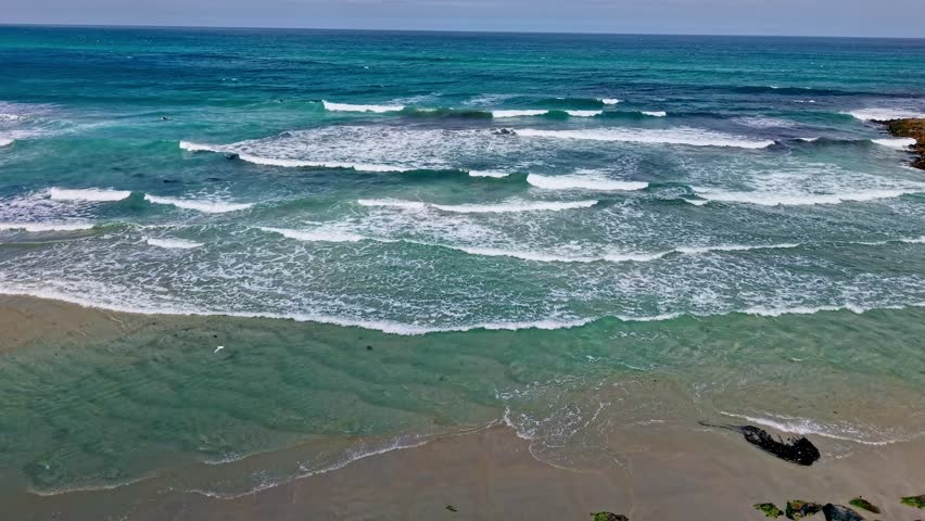 Drone shot ascending over the turquoise waters and waves rolling onto the sandy shore at St Kilda Beach, Dunedin, with seabirds visible in the water.