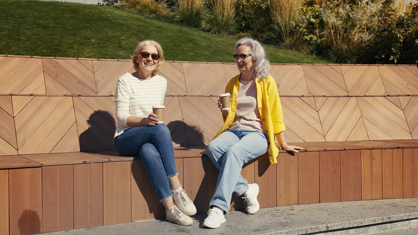 Two senior women drinking coffee and talking together on bench in city park