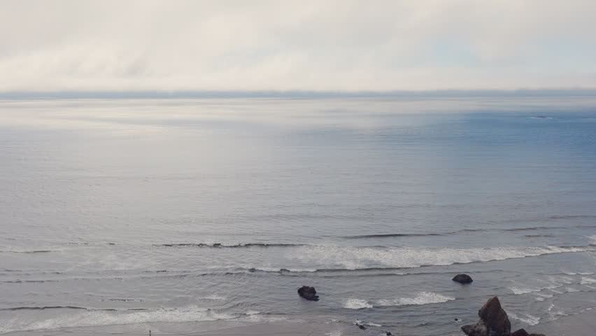 Cinematic aerial descending over the Pacific Ocean at Arcadia Beach on the Oregon Coast, revealing the shoreline and Douglas firs in the foreground on a calm, beautiful day.