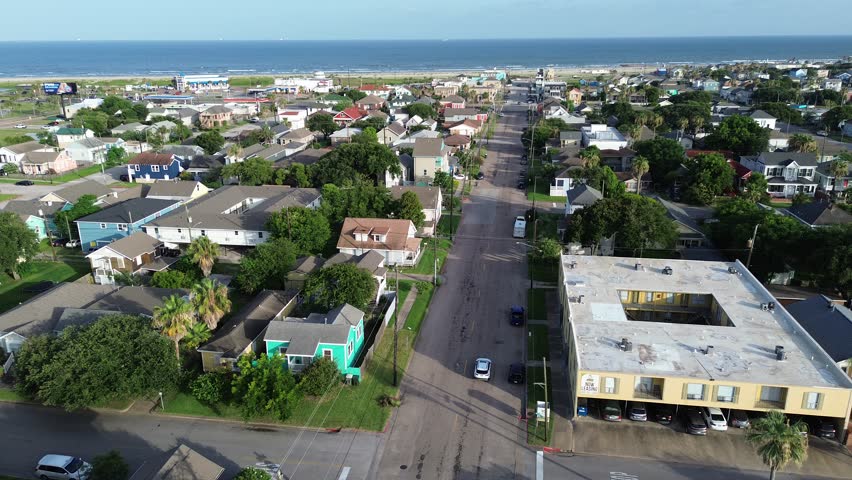 Classic Galveston residential neighborhood with colorful, elevated homes along deep green trees lines perpendicular street leading toward sandy shoreline of Stewart Beach area, Texas Gulf Coast. USA