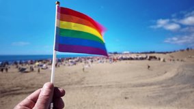 Person's hand holding a small waving LGBT rainbow pride flag in slow motion with a crowded beach in the background during the Winter Pride celebration in Maspalomas, Gran Canaria - Powered by Shutterstock - Get 15% off with code: PIKWIZARD15