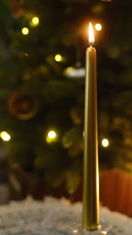 Vertical video. Golden candle glowing against background of Christmas tree. Winter holidays, cozy home concept.