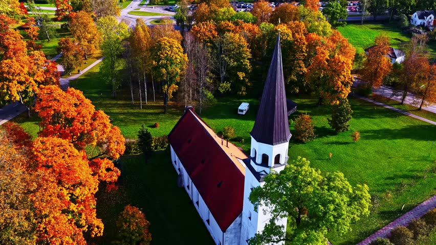 A circling pan shot orbits a white church with black steeple amid fiery fall foliage, revealing paths and rural serenity from above.