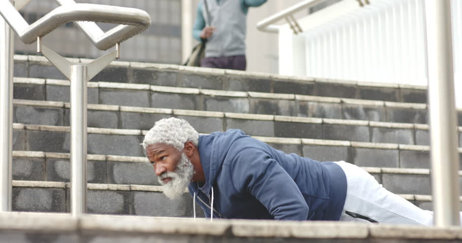 Starting push-ups on public stairway, senior African American man wearing wired earbuds for fitness. Fitness, strength, determination, urban, active, health, motivation