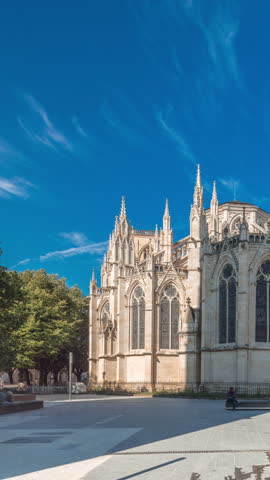Cathedrale Saint-Andre de Bordeaux timelapse hyperlapse with twin spires under a vibrant blue sky. Green trees and people walking in the square create a serene urban atmosphere. Bordeaux, France