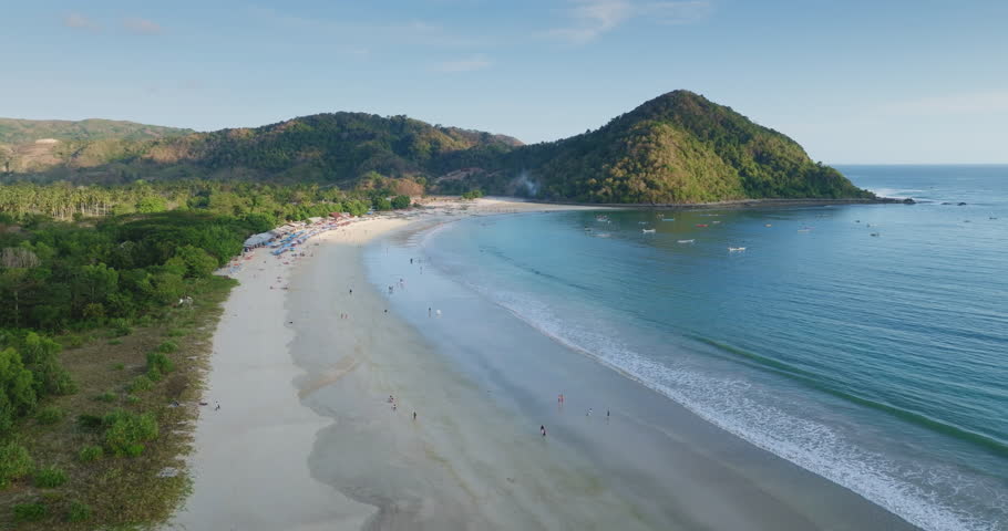 White sand beach on Selong Belanak bay with blue ocean water, lush green hills, distant mountains, and traditional fishing boats, attracting tourists for relaxation and watersports