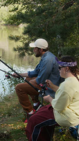 Vertical high angle shot of adult male and young woman with Down syndrome unwinding and casting their lines by waters edge in brightly lit forest while fishing