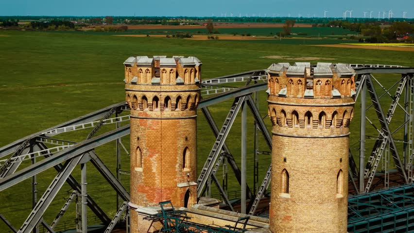 Steel railway bridge passing through twin brick towers over green fields. Metal truss crossing joining cylindrical fort like structures in open landscape. Iron rail line threading between medieval