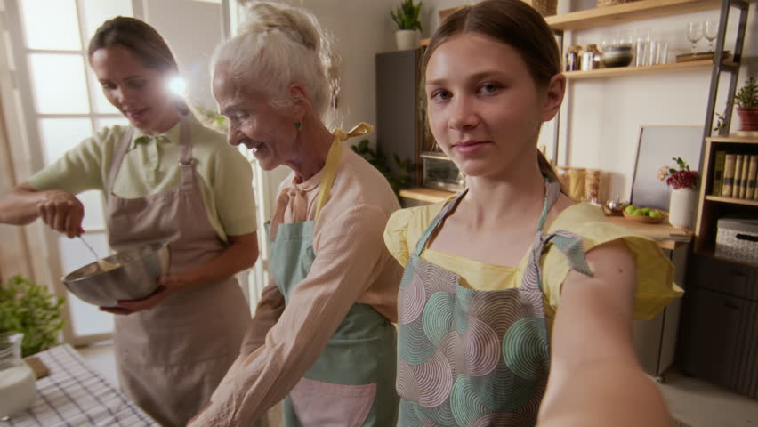 POV shot of smiling teenage girl taking selfie photo on mobile phone with mother and senior grandmother in aprons while baking biscuits in kitchen on weekend, slow motion