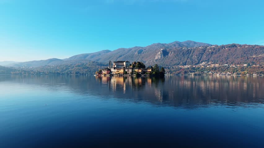 Aerial view of Isola San Giulio's buildings mirrored in the calm waters of Lake Orta, under a clear sky, creating a serene and picturesque scenery, Isola San Giulio, Piemonte, Italy.
