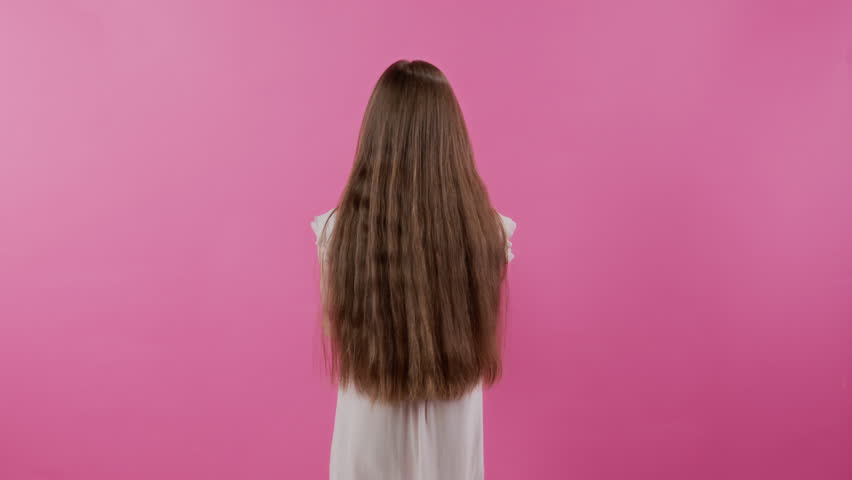 Studio portrait of playful teenage girl with long hair holding teddy bear up then turning to camera with joyful smile while posing on pink backdrop, concept of childhood