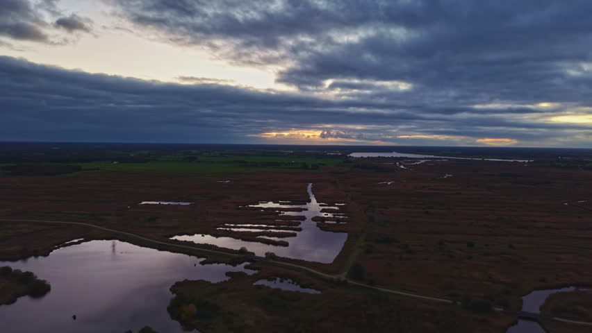 Golden hues merge with dark clouds as the sun sets over the tranquil wetlands, creating a peaceful scene of water and land under changing skies.