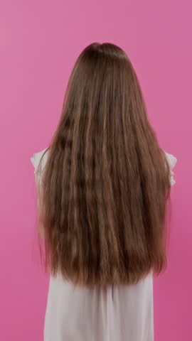 Vertical studio portrait of teenage girl with long hair playing with stuffed toy then turning to camera with smile while posing against pink background, childhood concept