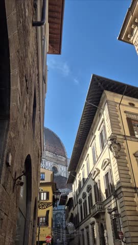 Vertical view of Brunelleschi dome of Florence Cathedral seen from a narrow historic street, framed by classic Renaissance buildings under a blue sky.